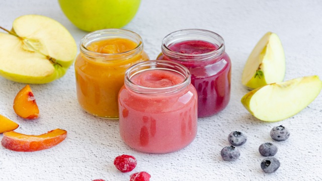 Three glass jars filled with colorful fruit puree—yellow, pink, and purple—surrounded by fresh apple slices, peach wedges, raspberries, and blueberries on a light textured surface